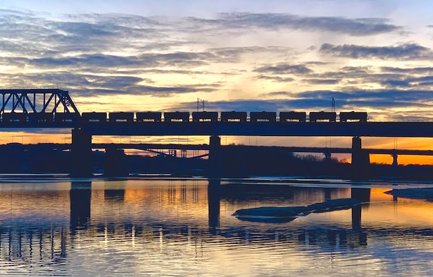 A long freight train traversing a modern railway bridge over a wide river.