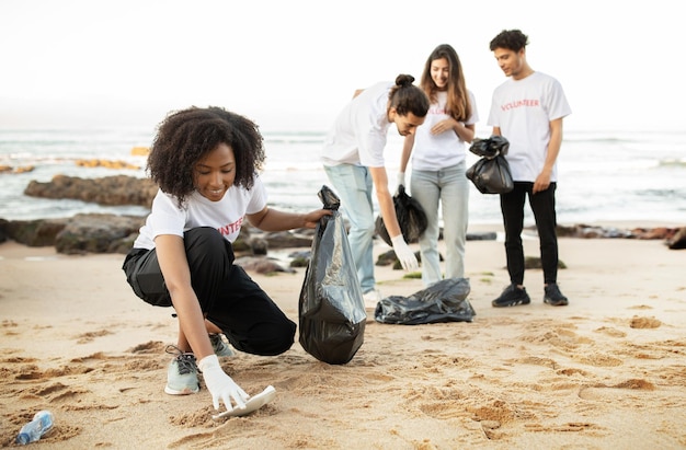 A group of high school students are participating in a beach cleanup, collecting trash and sorting recyclables. They are wearing gloves and carrying bags, with the ocean and a clear sky in the background, illustrating active participation in environmental preservation.