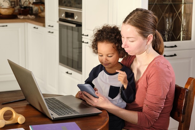 A photo of a teacher using a smartphone and laptop to teach a student remotely in Brazil during the pandemic. The setting could be a simple home environment, emphasizing the resourcefulness of the teacher. The focus is on the interaction and the effort to maintain education despite the challenges.