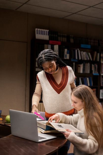 A medium shot of a teacher helping a young student in a public school library. The student is smiling and engaged, with books opened on the table. The library is colorful and well-stocked, suggesting a supportive learning environment.