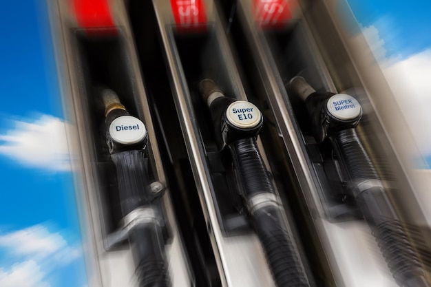 Close-up of a gas pump nozzle filling a car's fuel tank, showing the price per liter/gallon increasing rapidly. The background is blurred, focusing on the numbers changing on the pump.