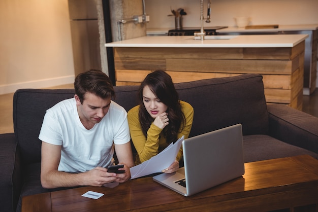 A young couple reviewing financial documents in their living room, possibly a mortgage statement, with a laptop displaying online banking information nearby.