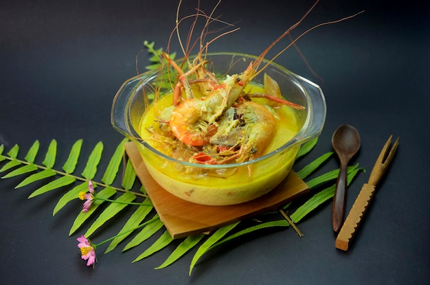 Close-up of a bowl of Tacacá, showing the tucupi broth, tapioca pearls, shrimp and jambu leaves. The bowl is set on a rustic wooden table with a woven placemat, and the background features blurred tropical foliage.